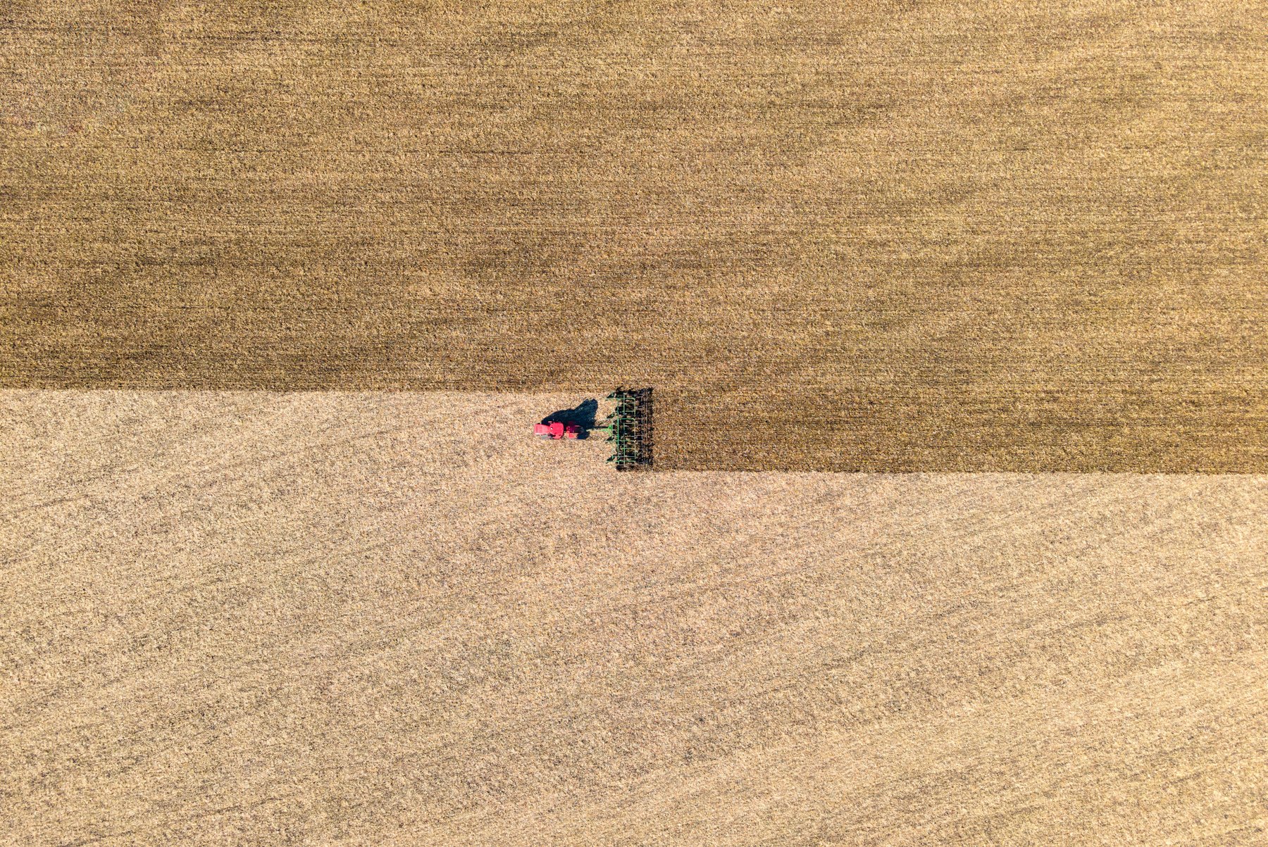 A tractor and implement working a pale field, seen from above, tiny in a very large empty plane.